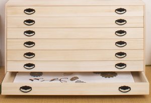 明るい色の木製チェストで、平らな水平の引き出しが 6 つ付いており、各引き出しには暗い色のハンドルが 2 つ付いています。一番下の引き出しが開いていると、中に平らな物が現れ、おそらくお手入れや虫干しのために保護された着物や書類が入っています。.
