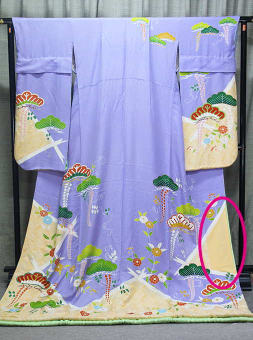 クリーム色の部分に色とりどりの花と松がデザインされた薄紫の引き振袖が、黒いラックに飾られている。着物の右下にピンクの楕円形の黄ばんだ部分がある。.